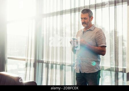 Senior man with beard holding mobile phone in hands working in airport business lounge while waiting for plane departure, sun flare from the window Stock Photo