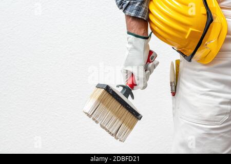 Caucasian house painter worker in white work overalls and with protective gloves, he holds the brush in his hand. Helmet and protective goggles. Work Stock Photo