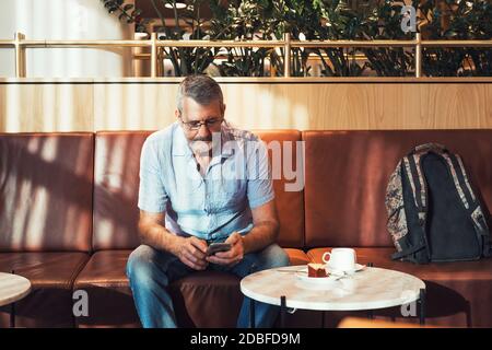 Senior man with beard holding mobile phone in hands working in airport business lounge while waiting for plane departure, sun flare from the window Stock Photo