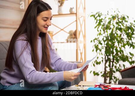 Comfortable. Woman opening, recieving greeting card for New Year and Christmas 2021 from friends or family. Reading a letter with best wishes, opening envelope. Holidays, celebration. Stock Photo