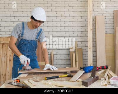 Asian carpenter with a hard hat and dustproof glasses use a sander machine to smooth plywood surfaces by abrasion with sandpaper. Morning work atmosph Stock Photo
