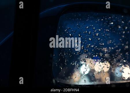 Car rearview mirror. Raindrops on the mirror reflecting the blurred lights of cars. Traffic and rainy weather on the road in the city. Light reflectio Stock Photo