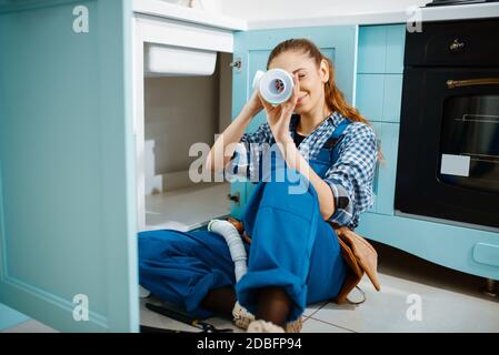 Female plumber in uniform lying on the floor in the kitchen, top view. Handywoman with toolbag repair sink, sanitary equipment service at home Stock Photo