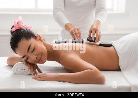 Relaxed asian woman enjoying hot stones back massage in spa salon, beautiful korean lady with flower in hair having beauty treatment therapy from prof Stock Photo