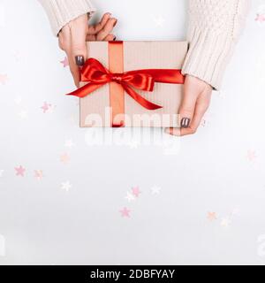 Womans hands holding cardboard present box with red bow on white background with multicolored star confetti. Square shape for social media Stock Photo