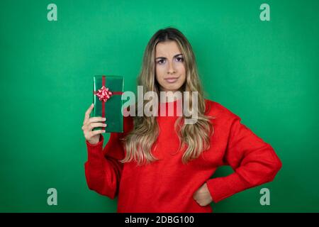 Young beautiful girl holding gift over isolated green background skeptic and nervous, disapproving expression on face with arms in waist Stock Photo