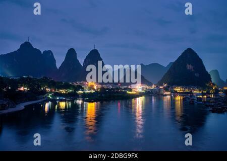 View of Yangshuo town illuminated in the evening with dramatic karst mountain landscape in background over Li river. Yangshuo, China Stock Photo