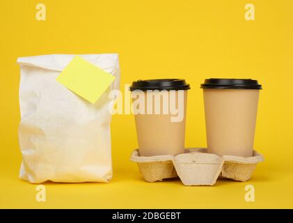 white paper bag, brown paper disposable cups with a plastic lid stand in the tray on a yellow background, takeaway containers Stock Photo