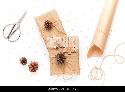 Gift box in craft paper, decorated with a pine cone on a white background, scissors and wrapping paper nearby. The concept of preparing gifts for the holiday Stock Photo