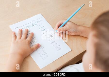 Child solving maths exercises. 7 years old boy doing maths lessons sitting at desk in his room. View from his back, close up Stock Photo