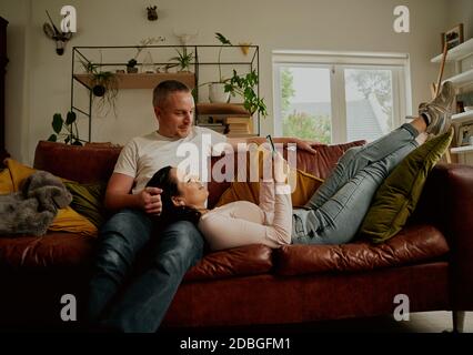 Beautiful young girlfriend using phone while relaxing on boyfriend lap on couch at home Stock Photo