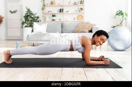 Beautiful black woman doing fitness training on sports mat at home, standing in elbow plank, strengthening abs muscles Stock Photo