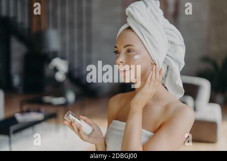 Beautiful young European woman with white bath towel on head, applies face cream and body lotion, enjoys skin care procedures after shower, poses at h Stock Photo