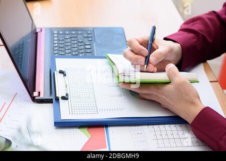 a person writes in a notebook data from a laptop and documents laid out on the table Stock Photo
