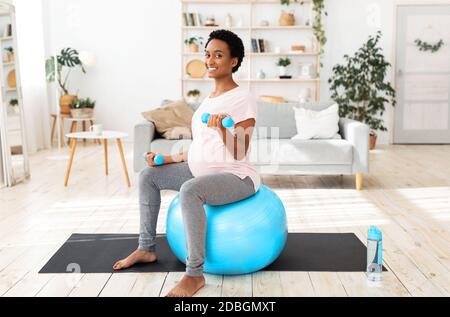 Happy black pregnant woman working out with dumbbells on fitball at home Stock Photo