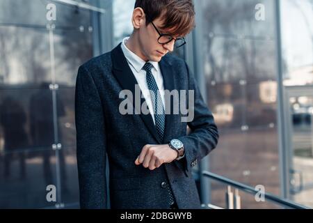 Stylish handsome young businessman in a business suit, late for work looks at the clock on his hand, standing against the background of a glass buildi Stock Photo