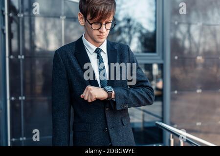 Stylish handsome young businessman in a business suit, late for work looks at the clock on his hand, standing against the background of a glass buildi Stock Photo