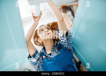 Female plumber in uniform lying on the floor in the kitchen, top view. Handywoman with toolbag repair sink, sanitary equipment service at home Stock Photo