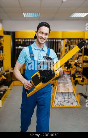Male worker in uniform holds chainsaw in tool store. Choice of professional equipment in hardware shop, electrical instrument supermarket Stock Photo