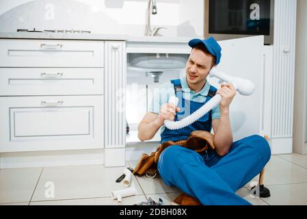 Male plumber in uniform listerns to the drain pipe in the kitchen, humor. Handywoman with toolbag repair sink, sanitary equipment service at home Stock Photo
