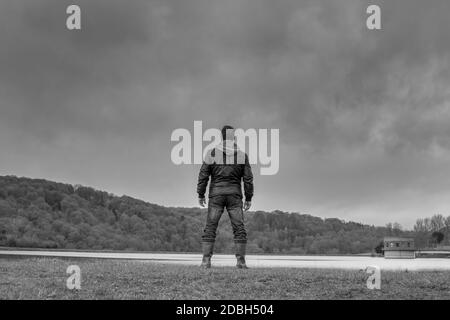 Black and white rear view of a menacing man with a hood looking over water with moody clouds above Stock Photo