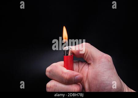 European white mans hand holds a burning red ignited plastic gas lighter with fire on an isolated black background Stock Photo