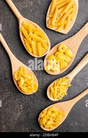 Various types of raw italian pasta in wooden spoons on black table. Top view. Stock Photo