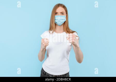 A young woman in a protective medical mask on her face shows a gesture of struggle and protection, on an isolated blue background. Air pollution, viru Stock Photo