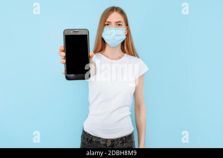 A young woman wearing a protective medical mask on her face shows a blank smartphone screen for a copy of the space, standing on an isolated blue back Stock Photo
