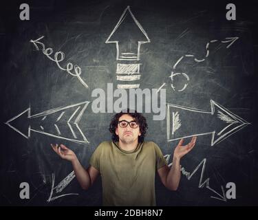 Perplexed man, hands outstretched, shrugging shoulders, has a problem with direction choice. Multiple different arrows sketches on the blackboard poin Stock Photo