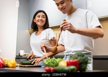 Happy Married Asian Couple Cooking Salad Together Standing In Kitchen Stock Photo