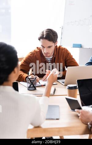 Focused office worker writing on notebook while sitting at desk with blurred african american woman gesturing on foreground Stock Photo