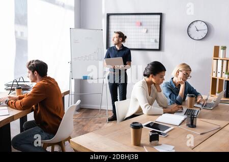 Young adult man with laptop looking away, while standing near multicultural co-workers sitting at workplaces in office Stock Photo