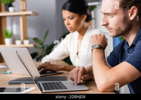 Focused office worker with watch using laptop while sitting at workplace with blurred african american woman on background Stock Photo