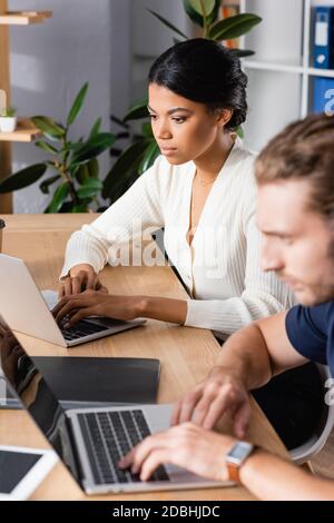 Focused african american woman looking at laptop while sitting at workplace with blurred man on foreground Stock Photo