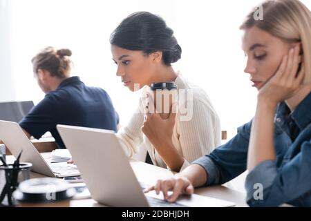 Focused african american woman with paper cup looking at laptop, while sitting at table with blurred tired woman on foreground Stock Photo