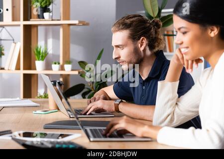 Focused young adult man looking at laptop while sitting at workplace with blurred african american woman on foreground Stock Photo
