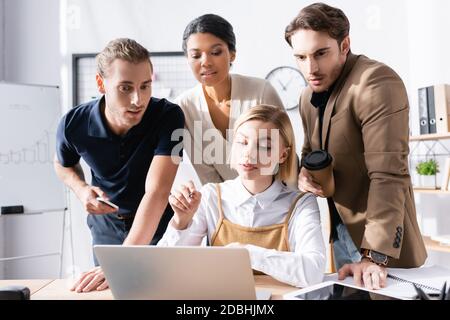 Focused woman pointing with pen, while sitting at table with focused multicultural office workers standing behind on blurred background Stock Photo