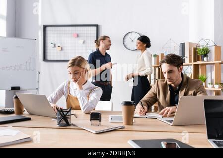 Bored office workers using laptop and writing on notebook while sitting at table, with multicultural colleagues talking on background Stock Photo