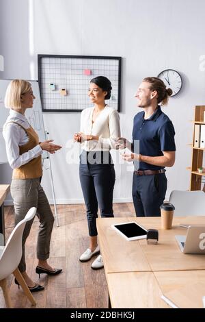 Smiling multicultural office workers talking while standing near workplace Stock Photo