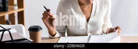 Cropped view of african american woman with pen sitting at desk with blank notebook in office on blurred background, banner