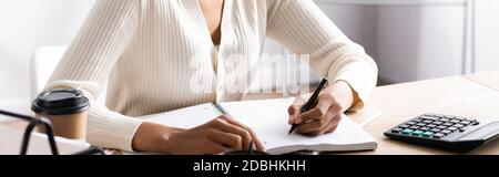 Cropped view of african american woman writing on blank notebook while sitting at desk on blurred background, banner