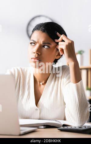 Thoughtful african american woman looking away while sitting at desk in office with blurred laptop on foreground Stock Photo