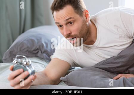Shocked Overslept Guy Holding Alarm Clock Lying In Bed Stock Photo