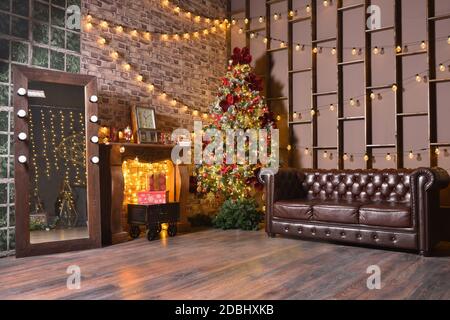 Stylish Christmas loft decorations in a brown living room with a leather sofa, Christmas tree and garlands with light bulbs on the wall Stock Photo