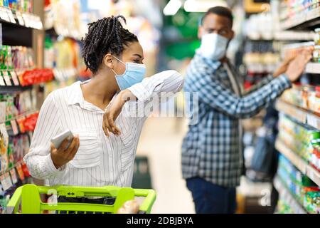 Sick African Woman Coughing In Elbow Wearing Mask In Supermarket Stock Photo