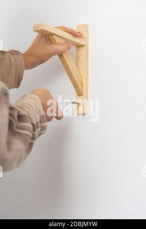 A man hangs a shelf in an apartment on a white wall, a worker fixes screws on a shelf corner with a screwdriver Stock Photo
