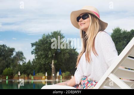 The sitting attractive blond hair woman is relaxing and enjoying in summer at poolside. Stock Photo