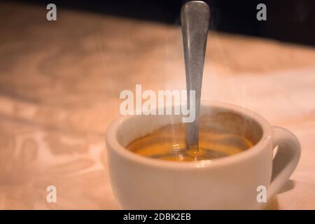 Smoke emitted from a cup with hot coffee and teaspoon in it. It placed on a tablecloth on a table. Close up with selective focus. Stock Photo