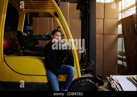 Inside the warehouse, the young woman businessman sits on the step of a yellow forklift and speaks on the phone. The sun is shining through the wareho Stock Photo